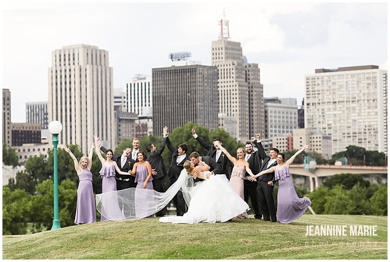 wedding party, bride, groom, kiss, dip, city backdrop, city skyline, green grass, hill, bridesmaids, groomsmen, cheers, arms up, Shepherd of the Lake Lutheran Church, The Great Hall, Hyatt Place, Instant Request DJ, Avant Decor, Linen Effects, Donatelli's, A Milestone Paper Co., Missy Cole, Unveil Loveliness, MIK Transportation, Eclipse Global Transportation, Jeannine Marie Photography, Minnesota wedding photographer, Saint Paul wedding photographer, summer wedding, lilac wedding, wedding inspiration, church wedding, Saint Paul wedding venues