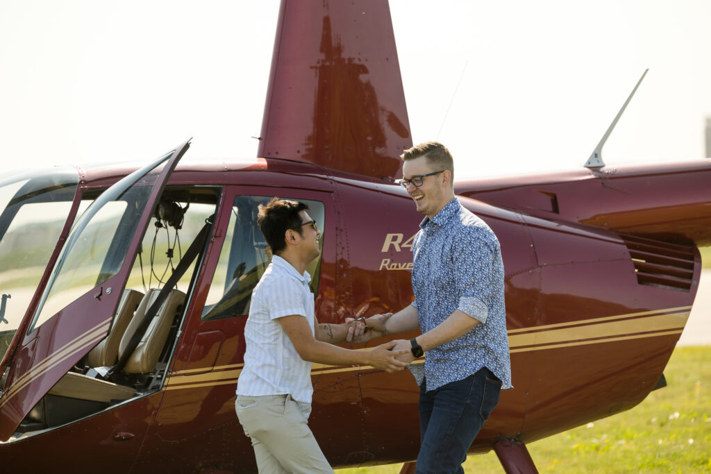 Gay couple consisting of a man with black hair and a taller man with blonde hair, standing hand in hand and looking at each other in front of a red helicopter after their wedding proposal at the Minneapolis-Saint Paul Airport.