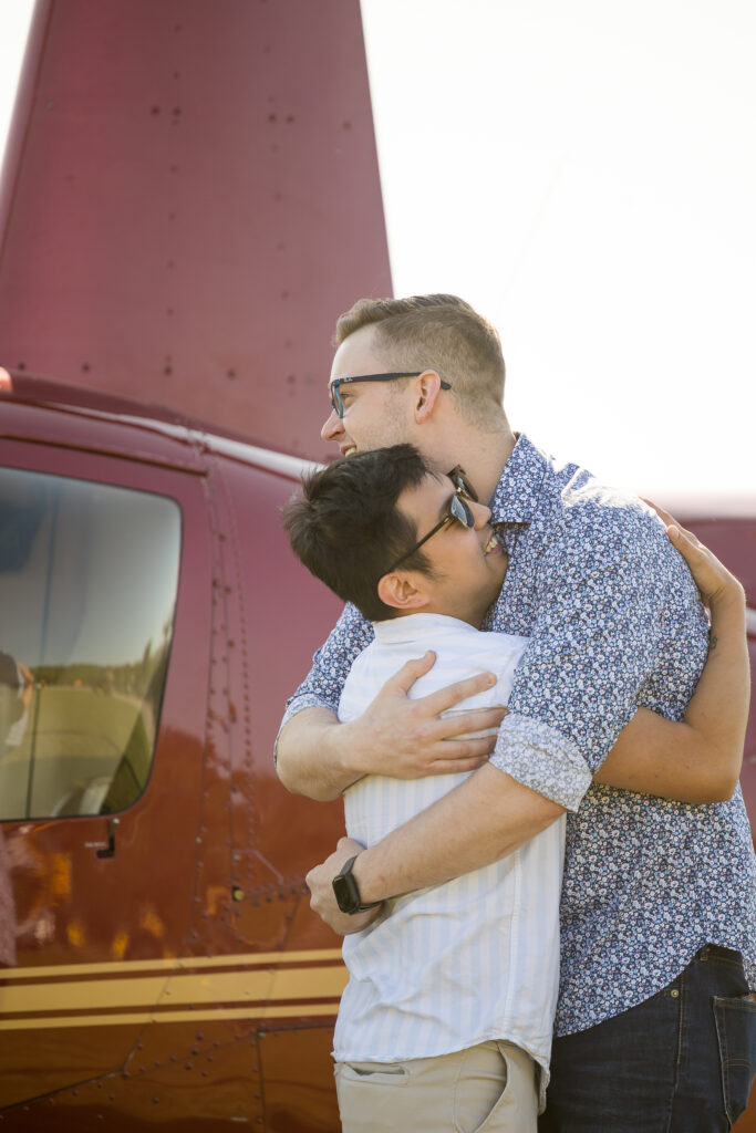 Unique wedding proposal photo of a gay couple embracing in front of a Red helicopter at Minnesota Helicopters in and Holman's Table in Saint Paul Minnesota, Taken by Saint Paul Photographer Jeannine Marie Photography.