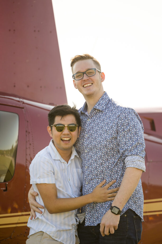 Gay couple posing with arms around one another. One is shorter and asiain decent while one is tall and caucasian. They are in front of a red Minnesota Helicopter's helicopter at Holman's Table in Saint Paul Minnesota after their Wedding Proposal captured by Jeannine Marie Photography.