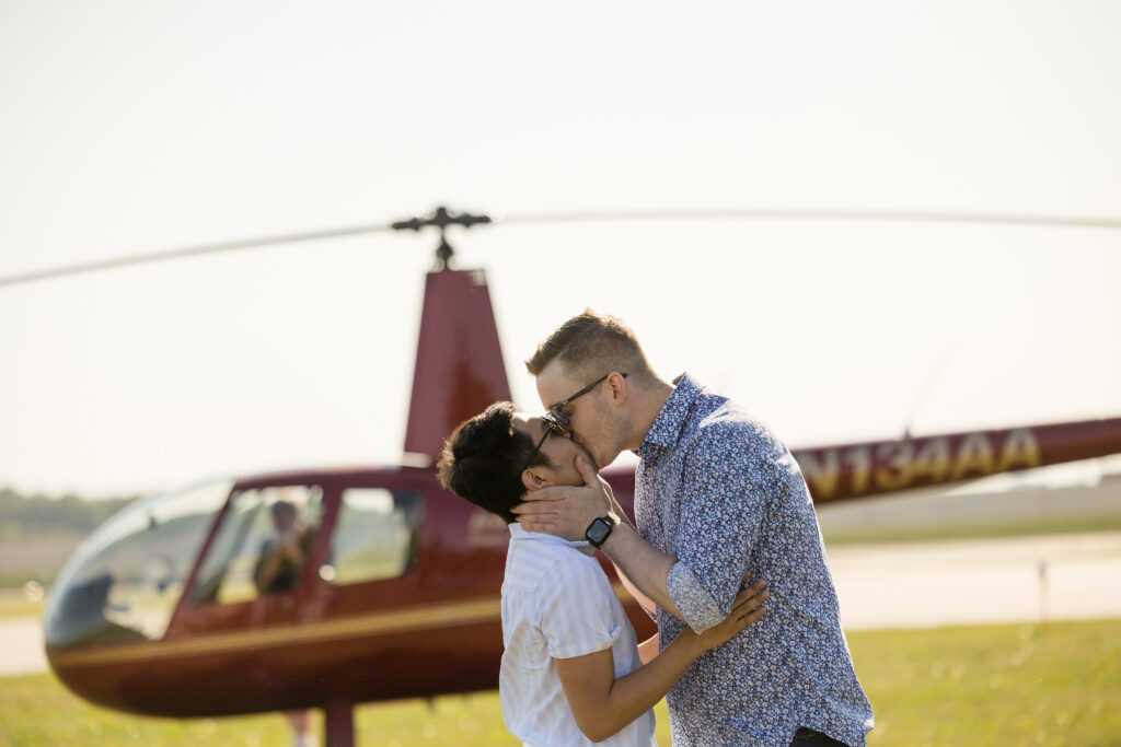 Gay couple consisting of a man with black hair and a taller man with blonde hair kissing each other with a red helicopter in the background at Holman's Table in Saint Paul Minnesota